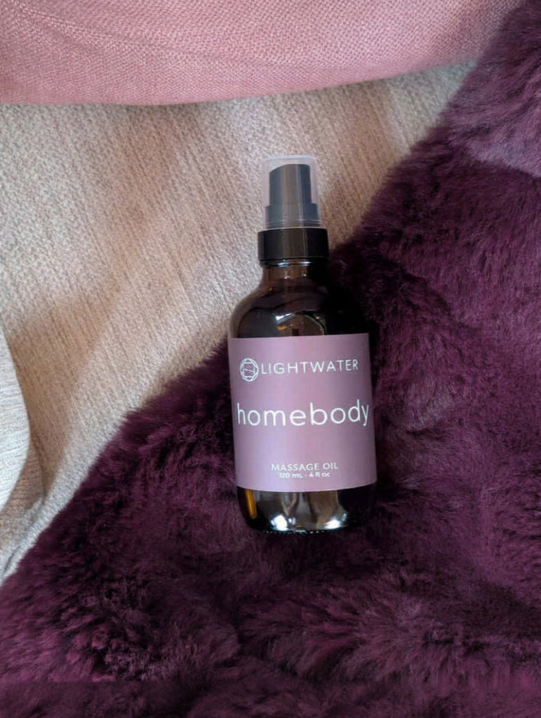A bottle of Lightwater Homebody Everyday Mist placed on a plush purple blanket with a cream knit fabric and pink fabric in the background.